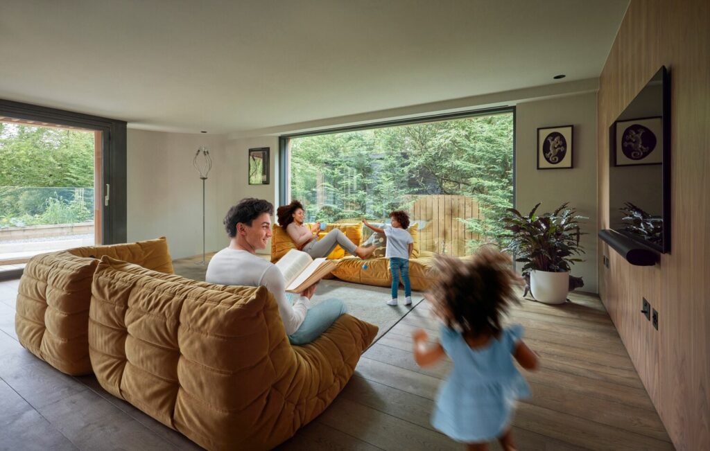 Famille dans un salon aux fauteuils moutarde, grande baie vitrée ouverte sur un jardin arboré
