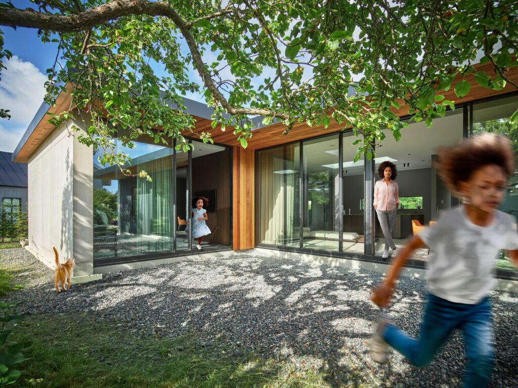 Une mère et ses deux filles traversent une cour gravillonnée entre deux volumes vitrés d'une maison contemporaine