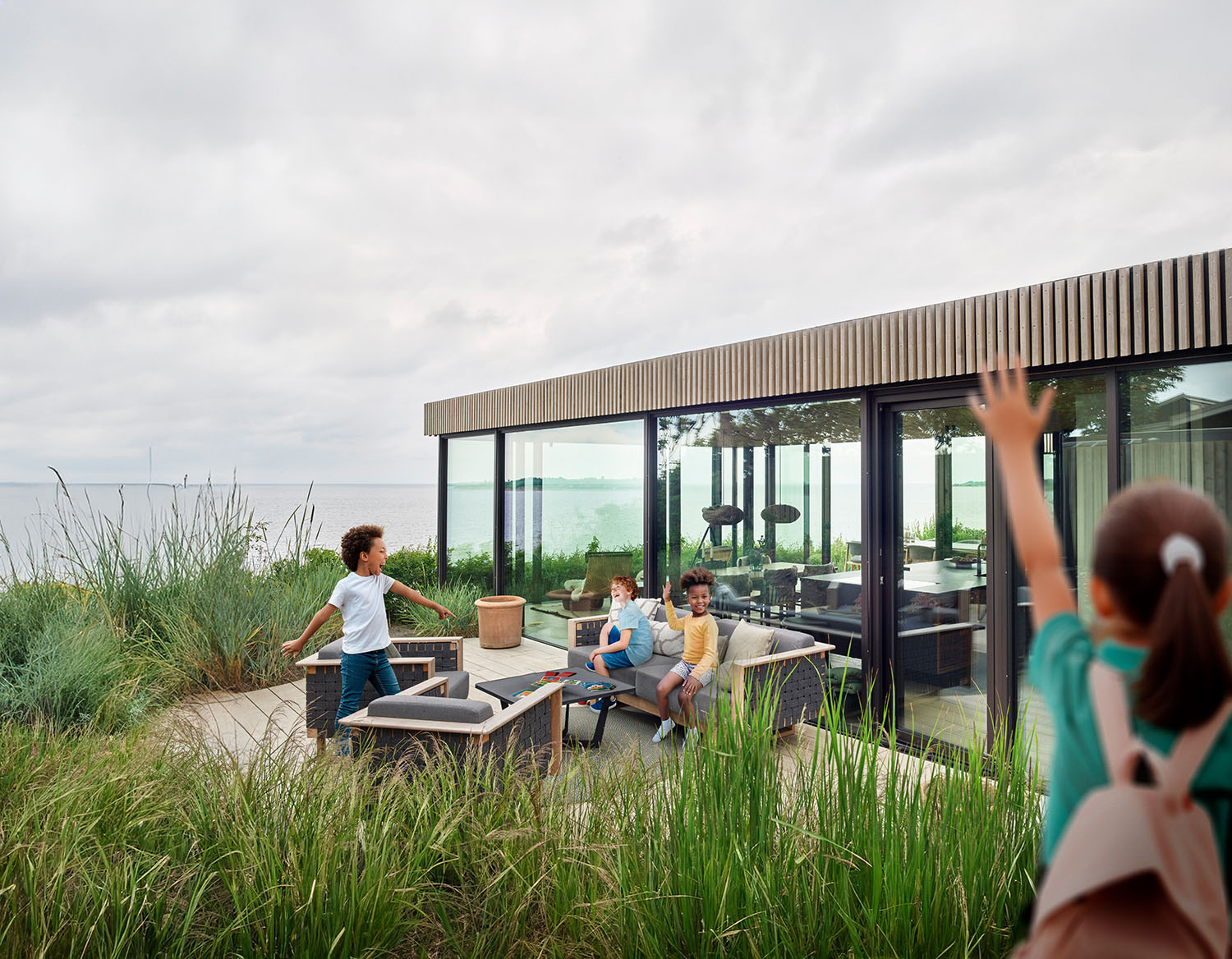 Enfants jouant sur une terrasse en bois face à la mer, devant une maison à larges baies vitrées